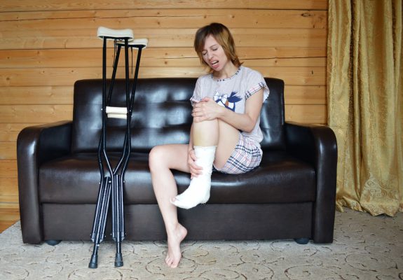 Mujer con la pierna escayolada sentada en un sofá junto a unas muletas