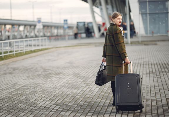 Viajera con abrigo y maleta de ruedas caminando hacia la terminal del aeropuerto